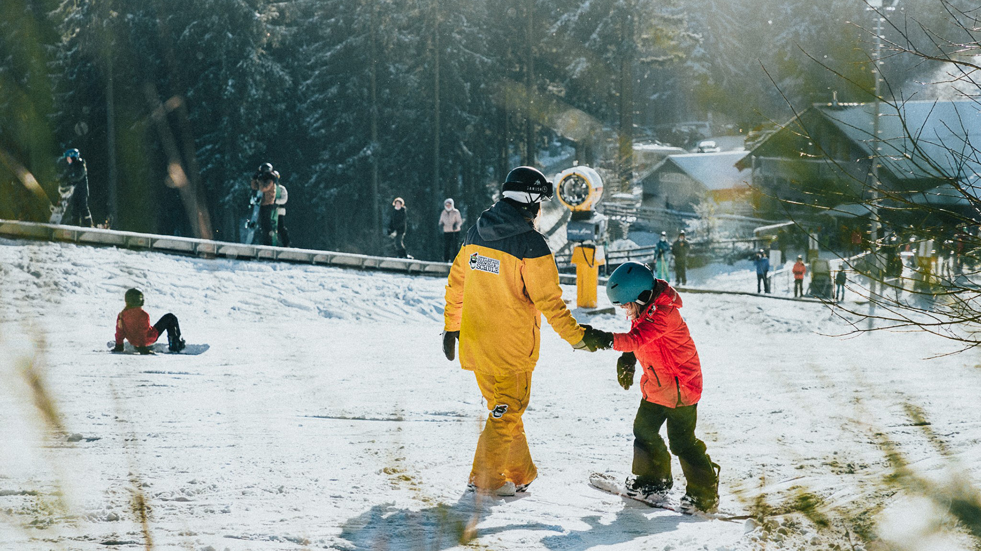 Ein Erwachsener hält ein auf einem Snowboard stehendes Kind an der Hand und hilft ihm beim Snowboard fahren lernen. Ein Erwachsener hält ein auf einem Snowboard stehendes Kind an der Hand und hilft ihm beim Snowboard fahren lernen.