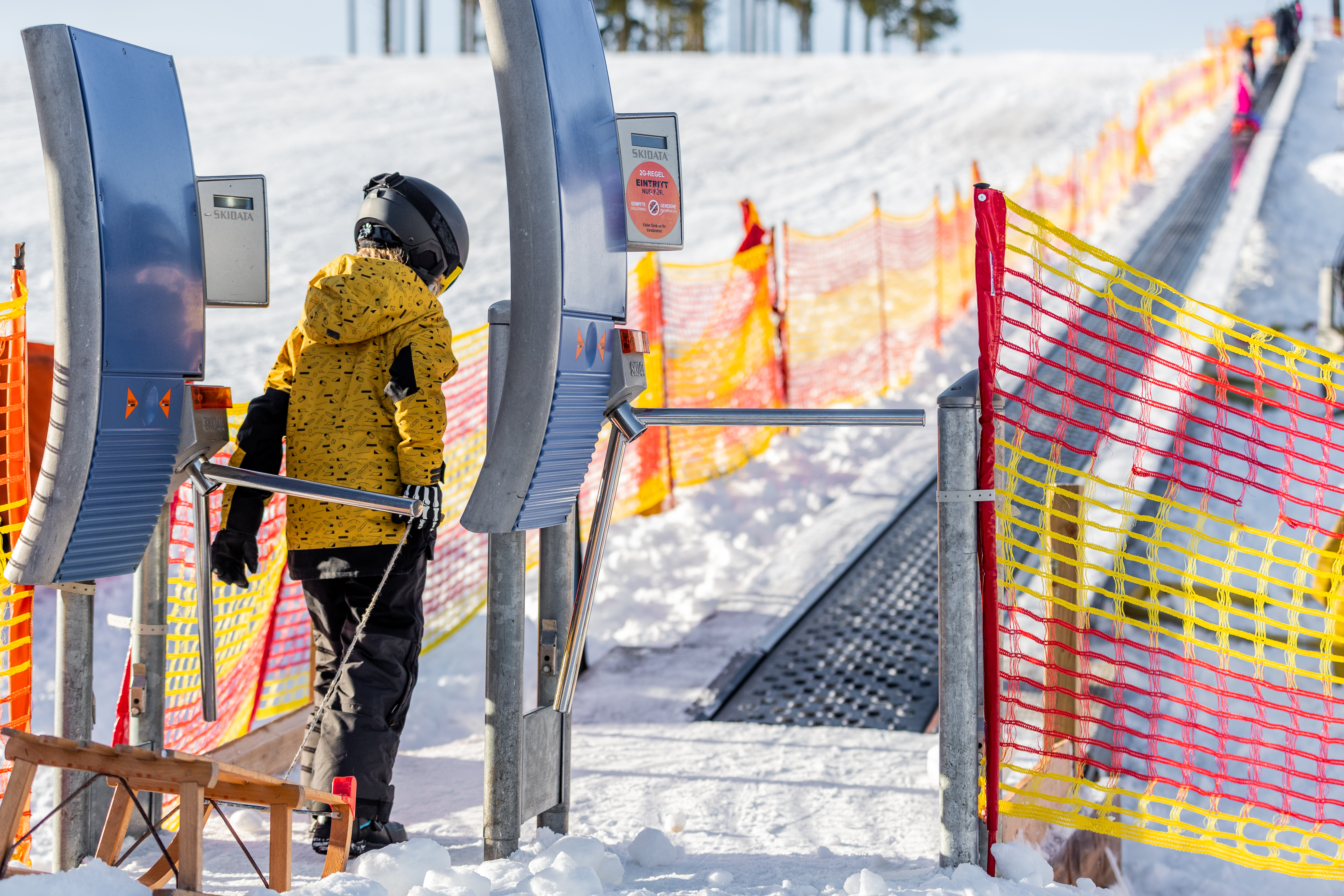 Kind geht mit Schlitten durch ein Drehkreuz zum Förderband.