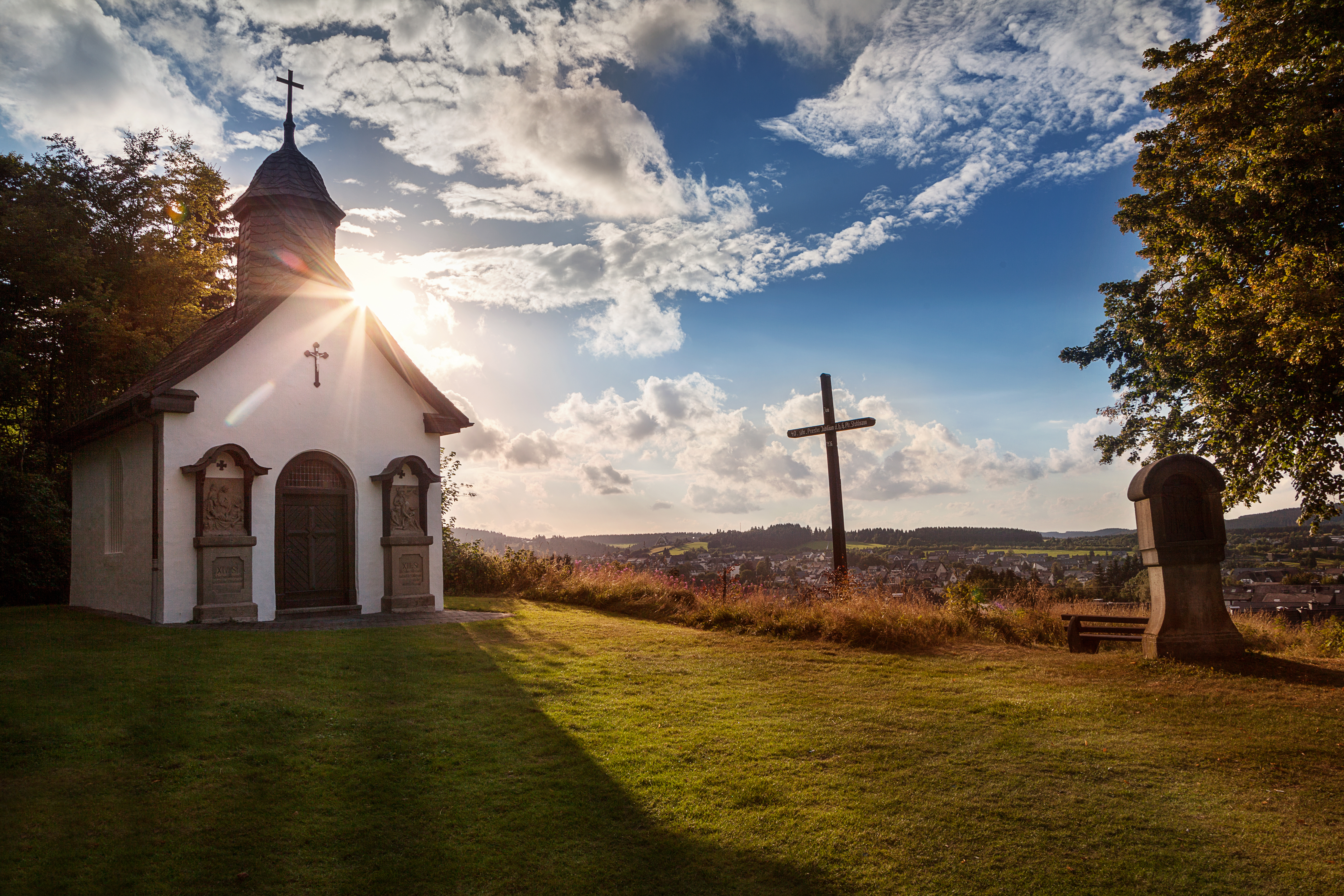 Eine Kapelle neben einem Kreuz und einer Kreuzwegstation mit blauem Himmel und der Sonne im Hintergrund.
