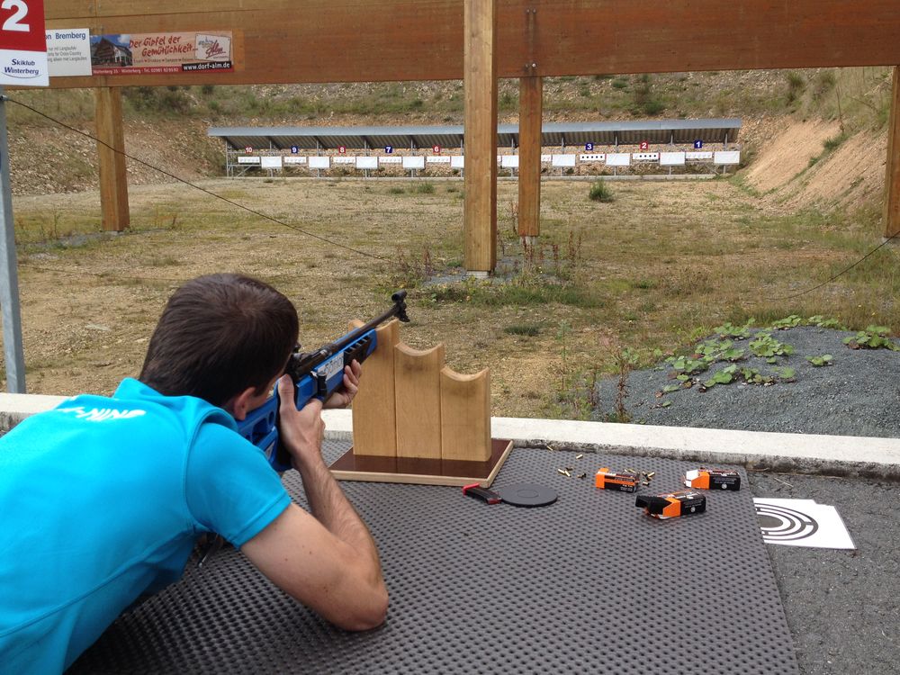 Ein auf dem Boden liegender Mann zielt mit einem Biathlongewehr auf eine Zielscheibe.