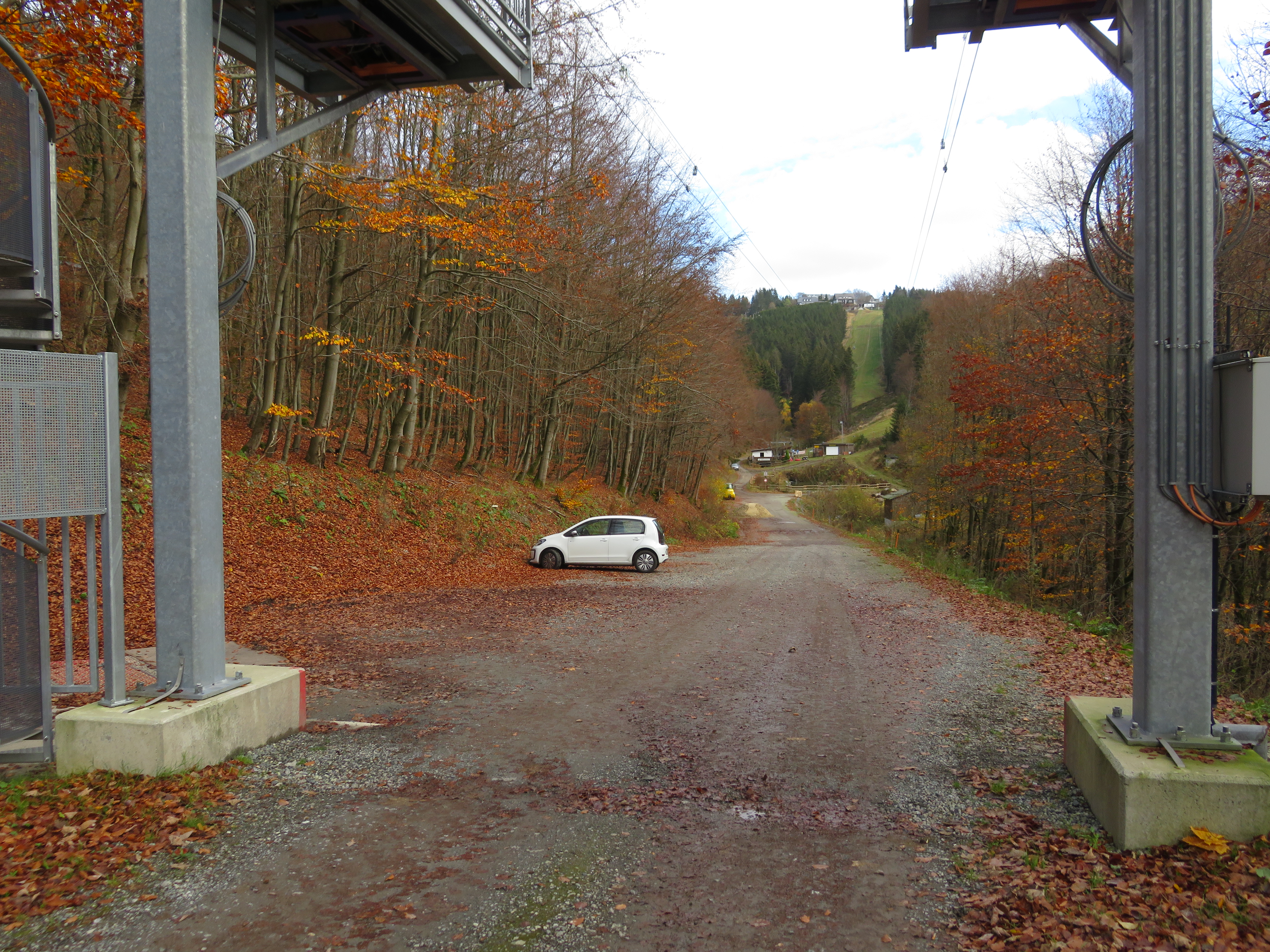 Parkplatz neben einem Waldweg mit einem weißen geparkten Auto, umgeben von Bäumen.