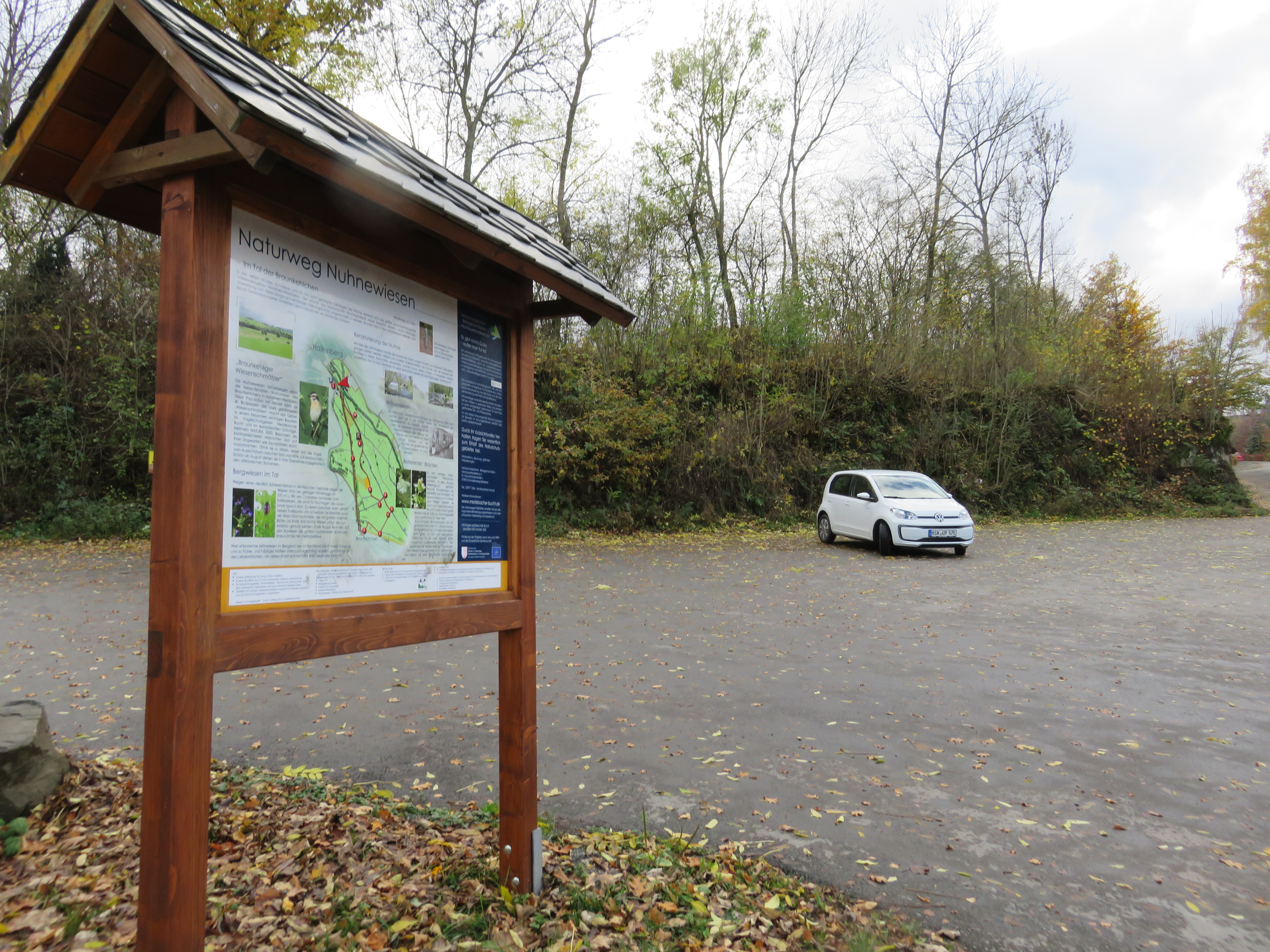 Parkplatz mit einem weißen geparkten Auto, im Vordergrund eine Informationstafel.