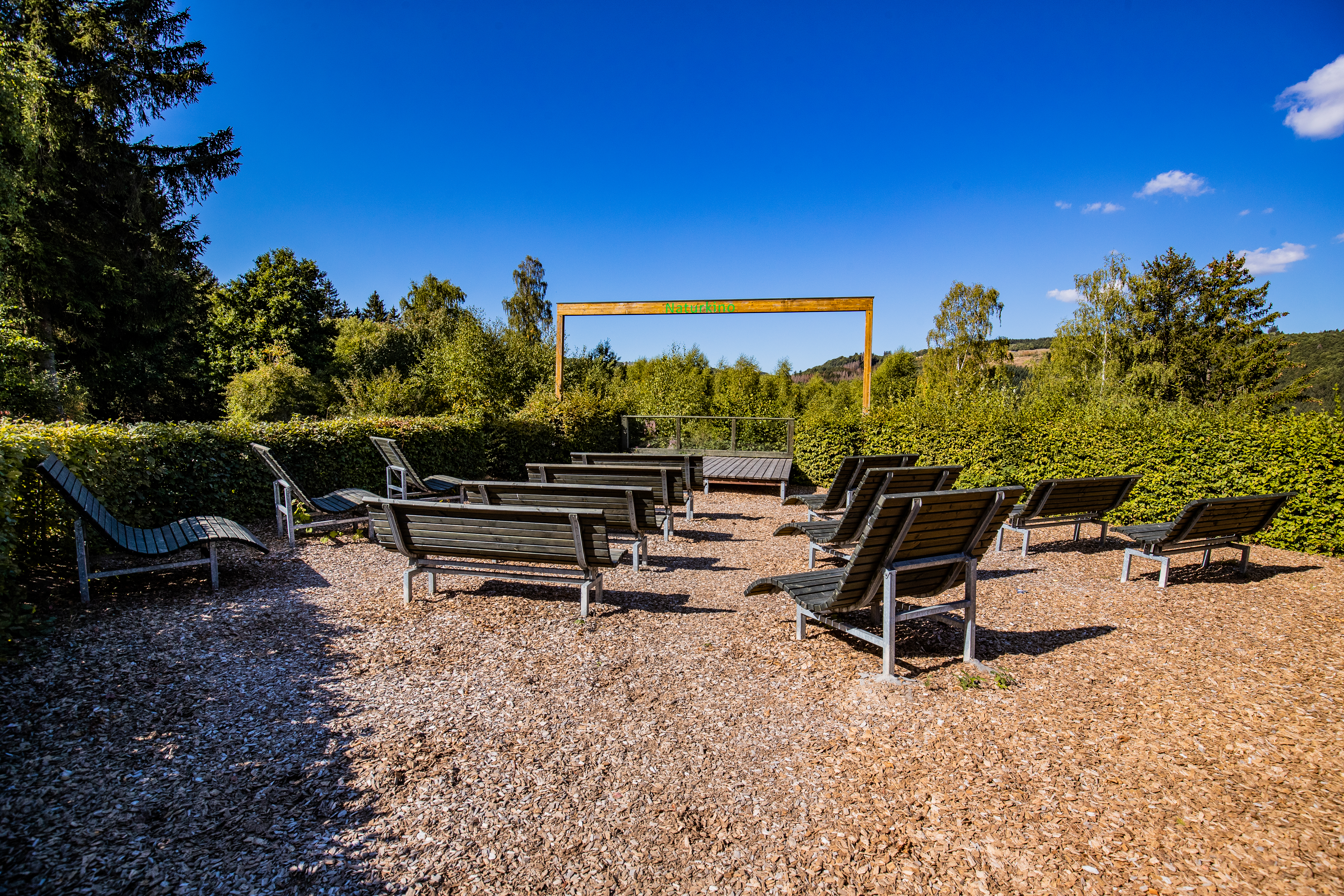 Einige Wellness-Sitzliegen vor einem hölzernen Rahmen umgeben von Bäumen und einer Hecke.