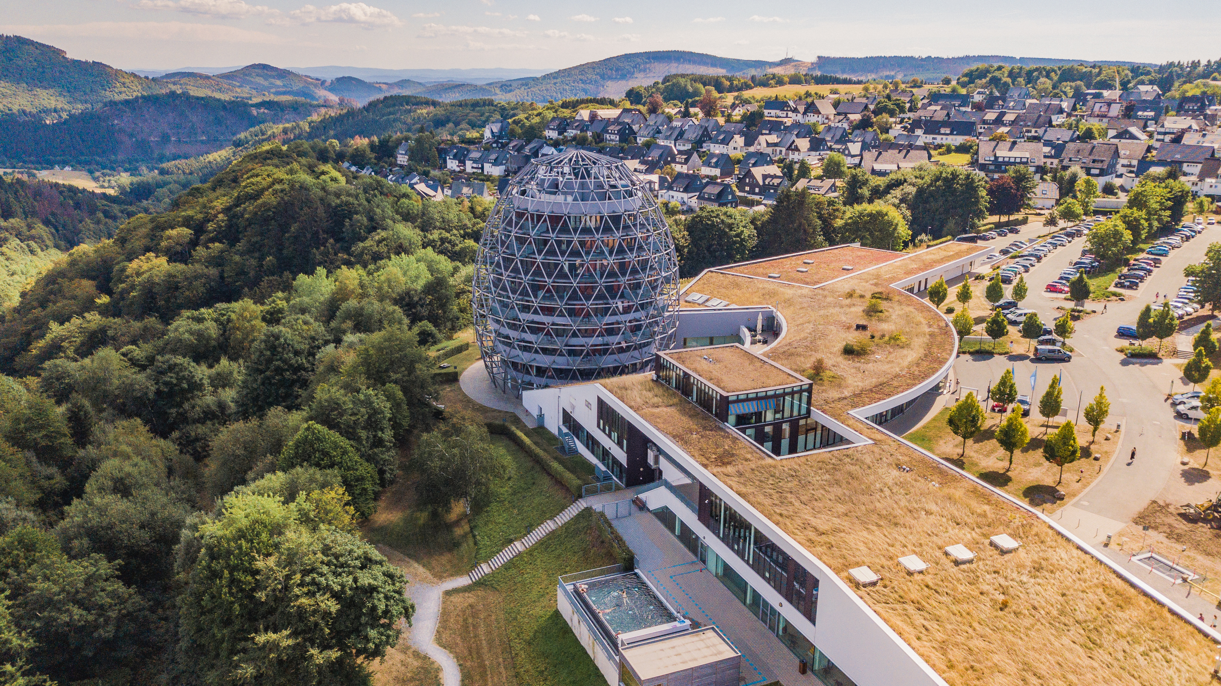 Drohnenaufnahme vom Hotel Oversum mit Parkplatz und Waldstück und einem Teil des Wohngebietes der Stadt Winterberg.