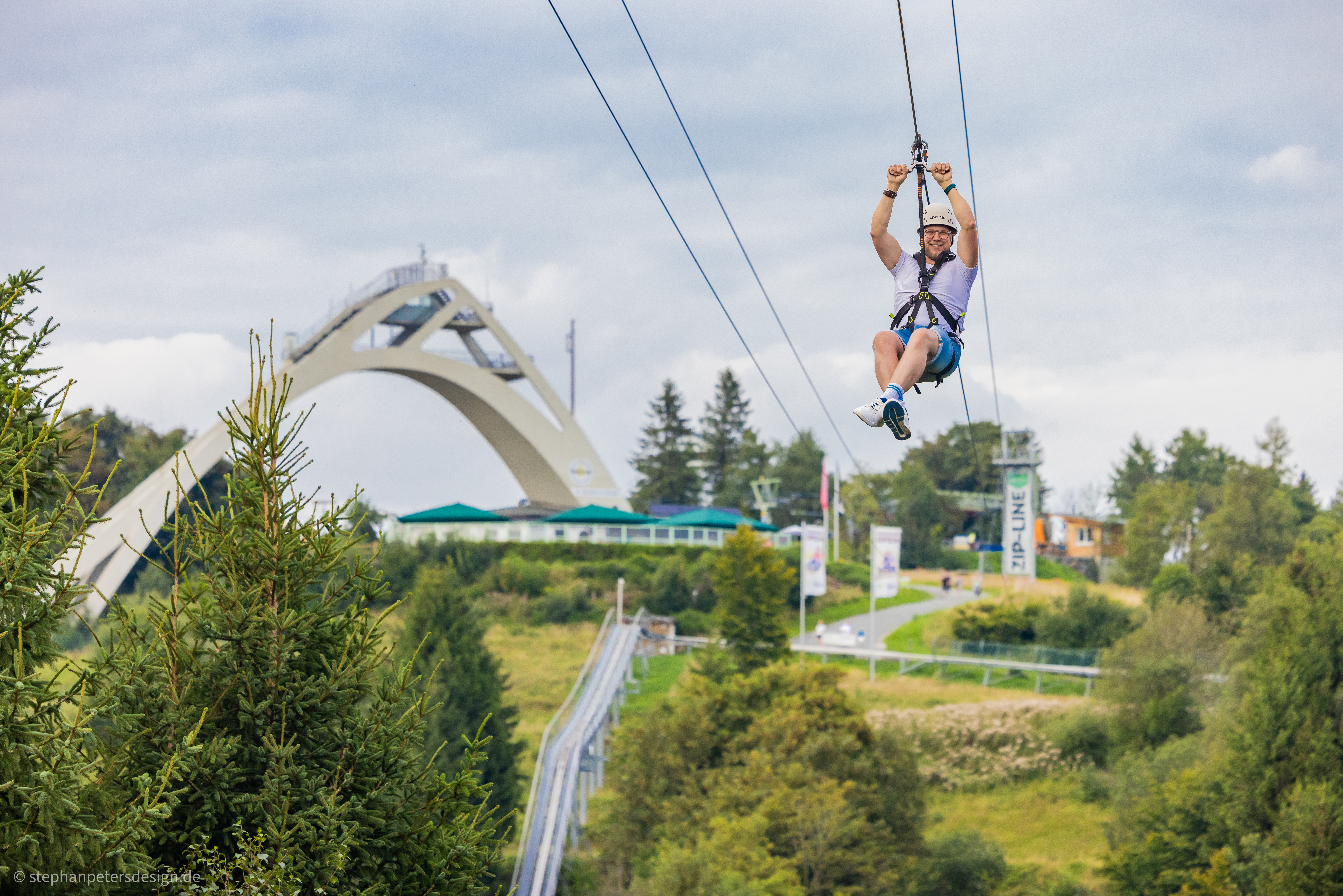 Die St. Georg Schanze umgeben von Bäumen und Wiesen, im Vordergrund ein Mann der an der Zipline fliegt.
