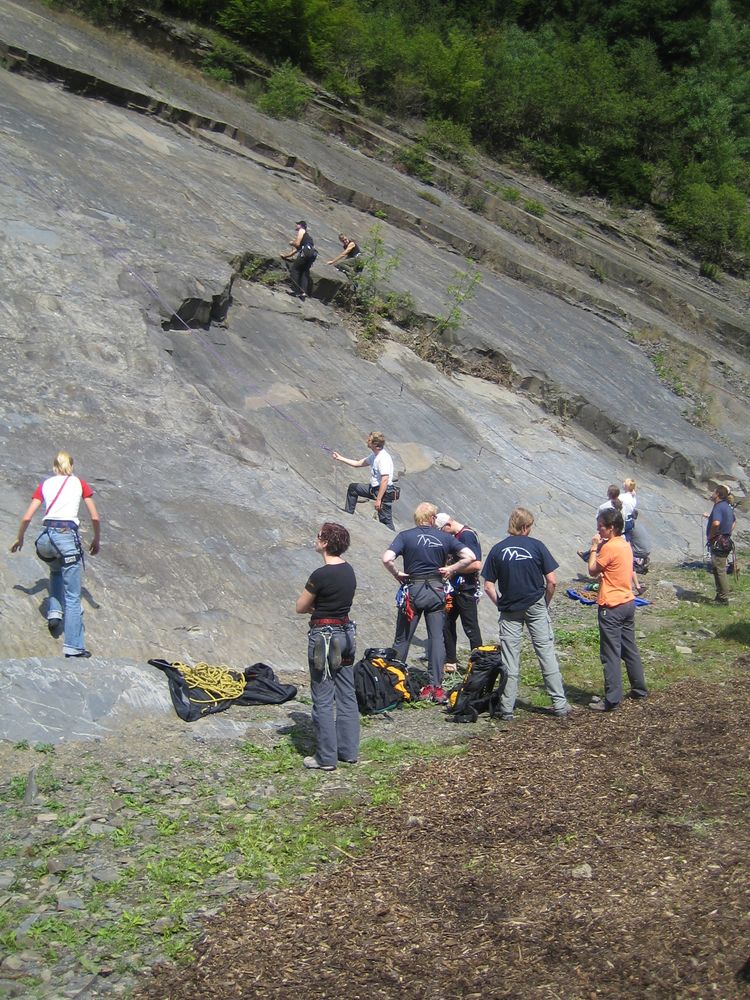 Einige Personen in Kletterausrüstung stehen vor einem Steinbruch und schauen einer Person beim Klettern zu. Einige Personen in Kletterausrüstung stehen vor einem Steinbruch und schauen einer Person beim Klettern zu.