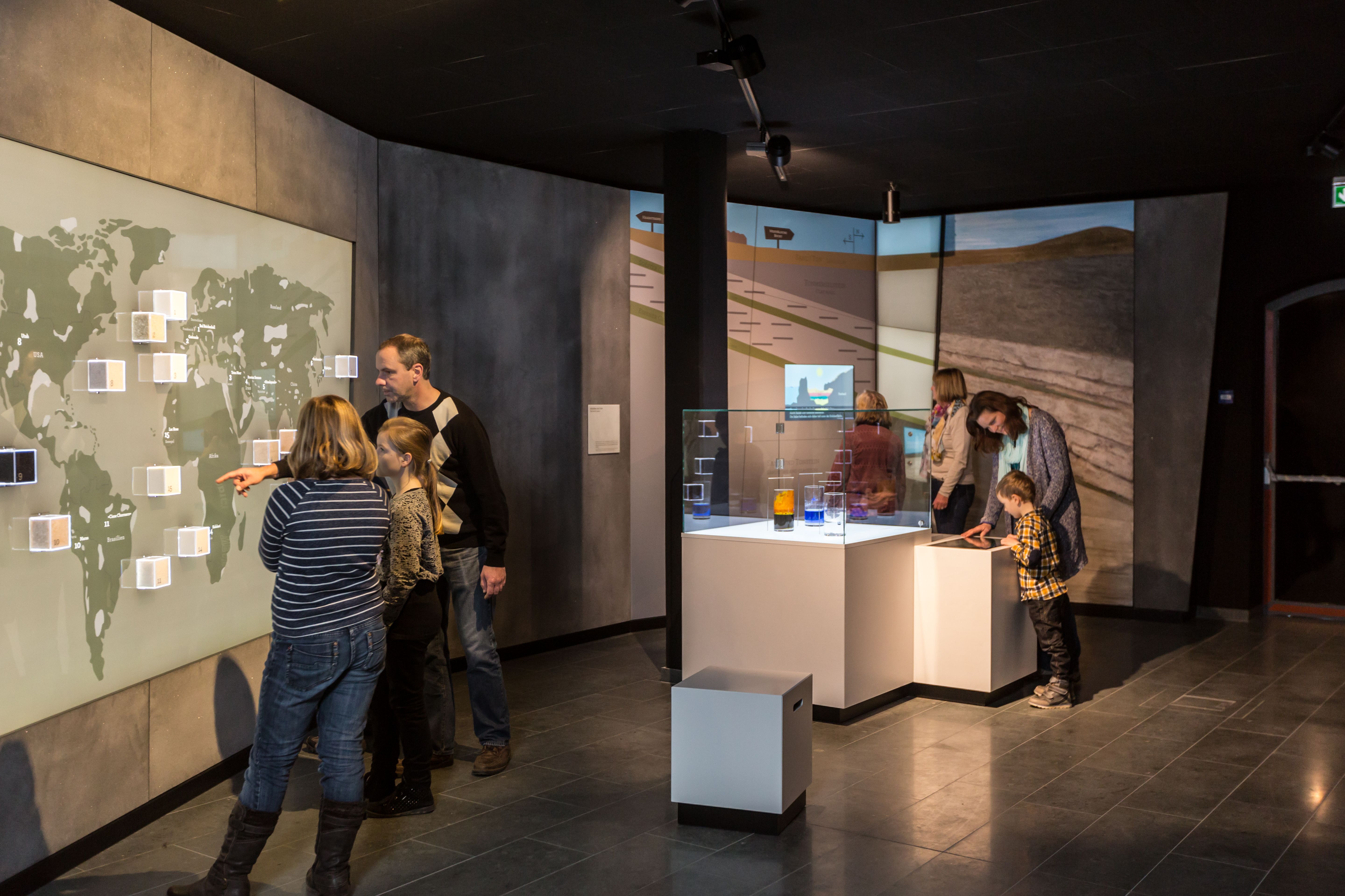 Ausstellungsraum mit Ausstellungsstücken in Glaskästen und einigen Personen, die eine Weltkarte an der Wand betrachten. Ausstellungsraum mit Ausstellungsstücken in Glaskästen und einigen Personen, die eine Weltkarte an der Wand betrachten.