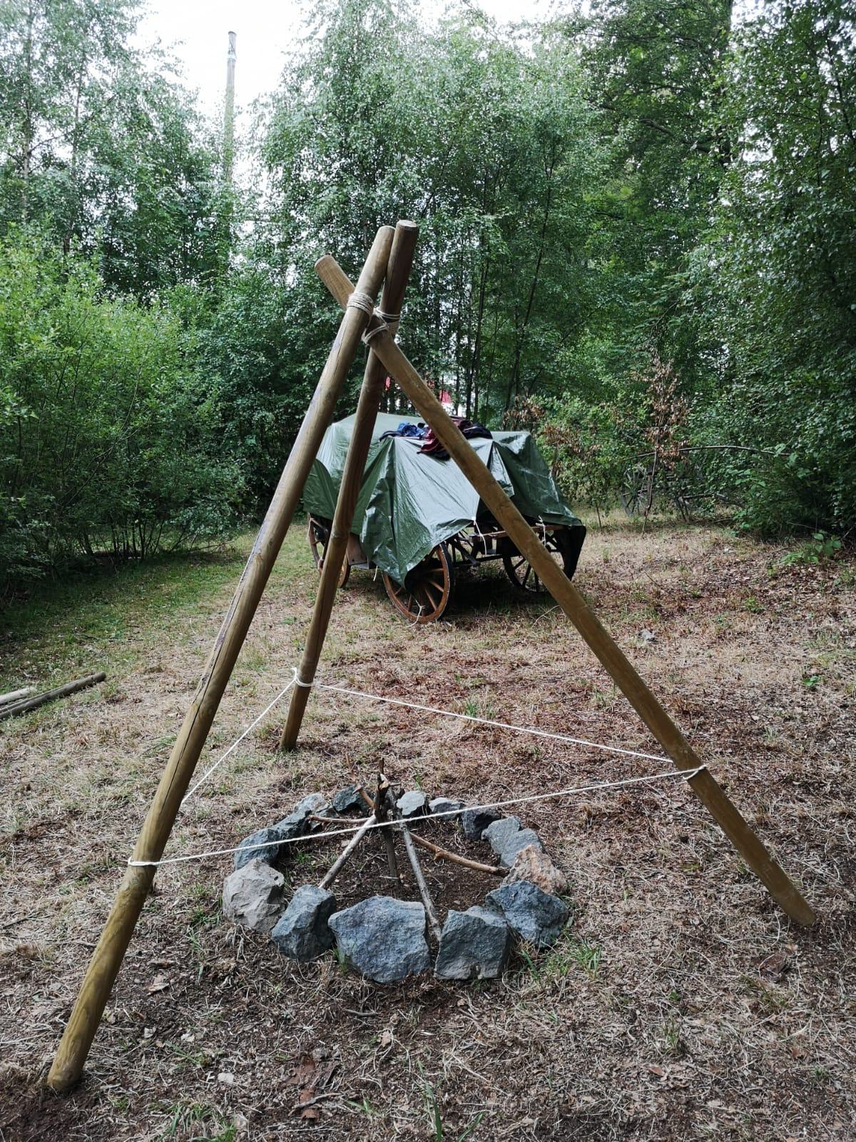 Eine Feuerstelle mit drei Holzstangen, die zu einem Zelt darüber aufgebaut sind und einer mit einer Folie abgedeckten Kutsche im Hintergrund.