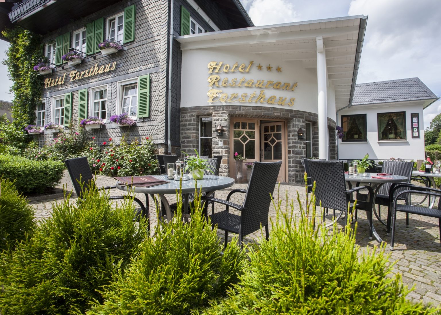 Blick über eine Terasse mit Sitzmöglichkeiten zu einem Gebäude mit der Aufschrift Hotel Restaurant Forsthaus. Blick über eine Terasse mit Sitzmöglichkeiten zu einem Gebäude mit der Aufschrift Hotel Restaurant Forsthaus.