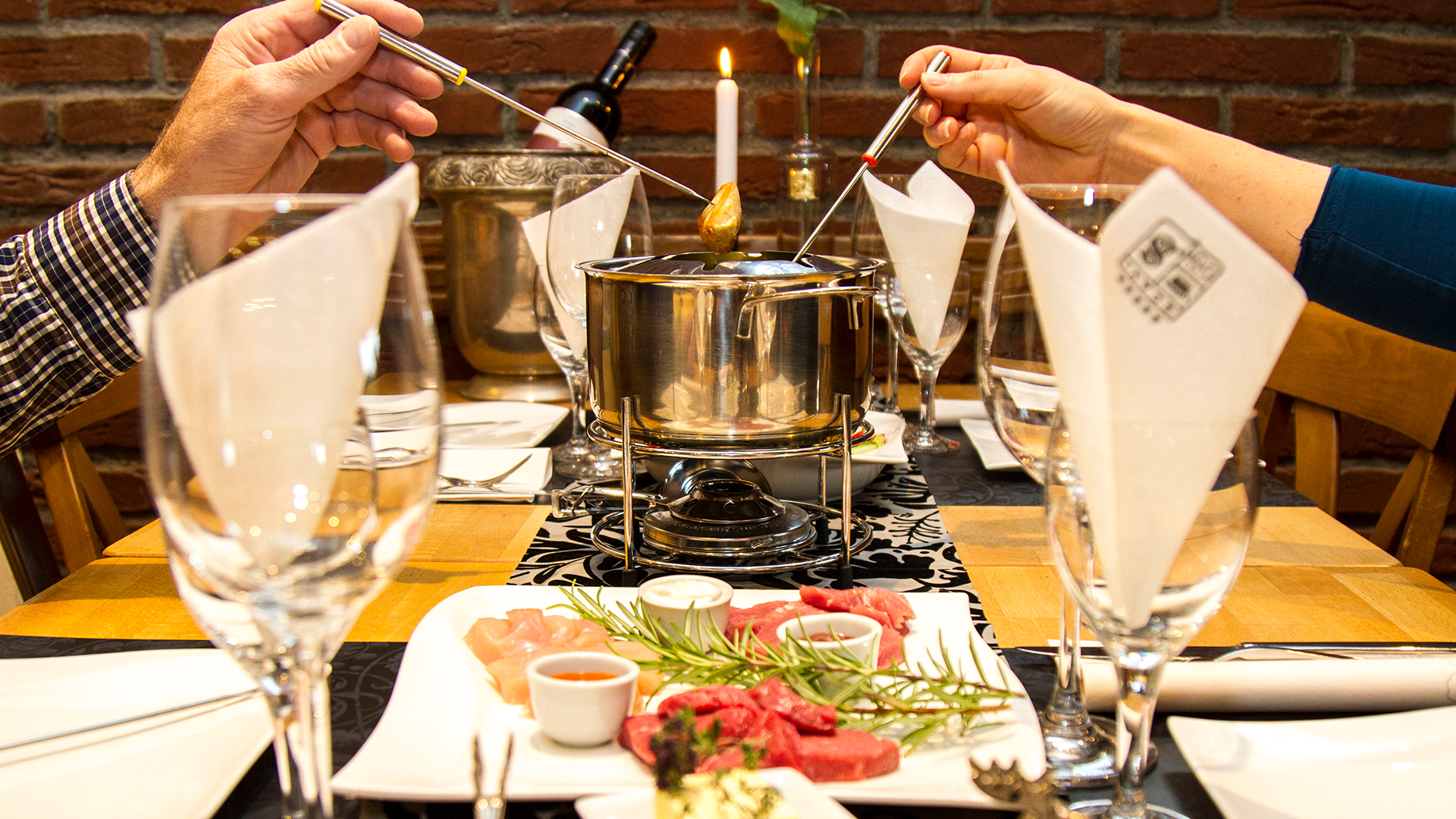 Ein eingedeckter Tisch mit zwei Händen, die lange Gabeln in einen Fonduetopf halten.