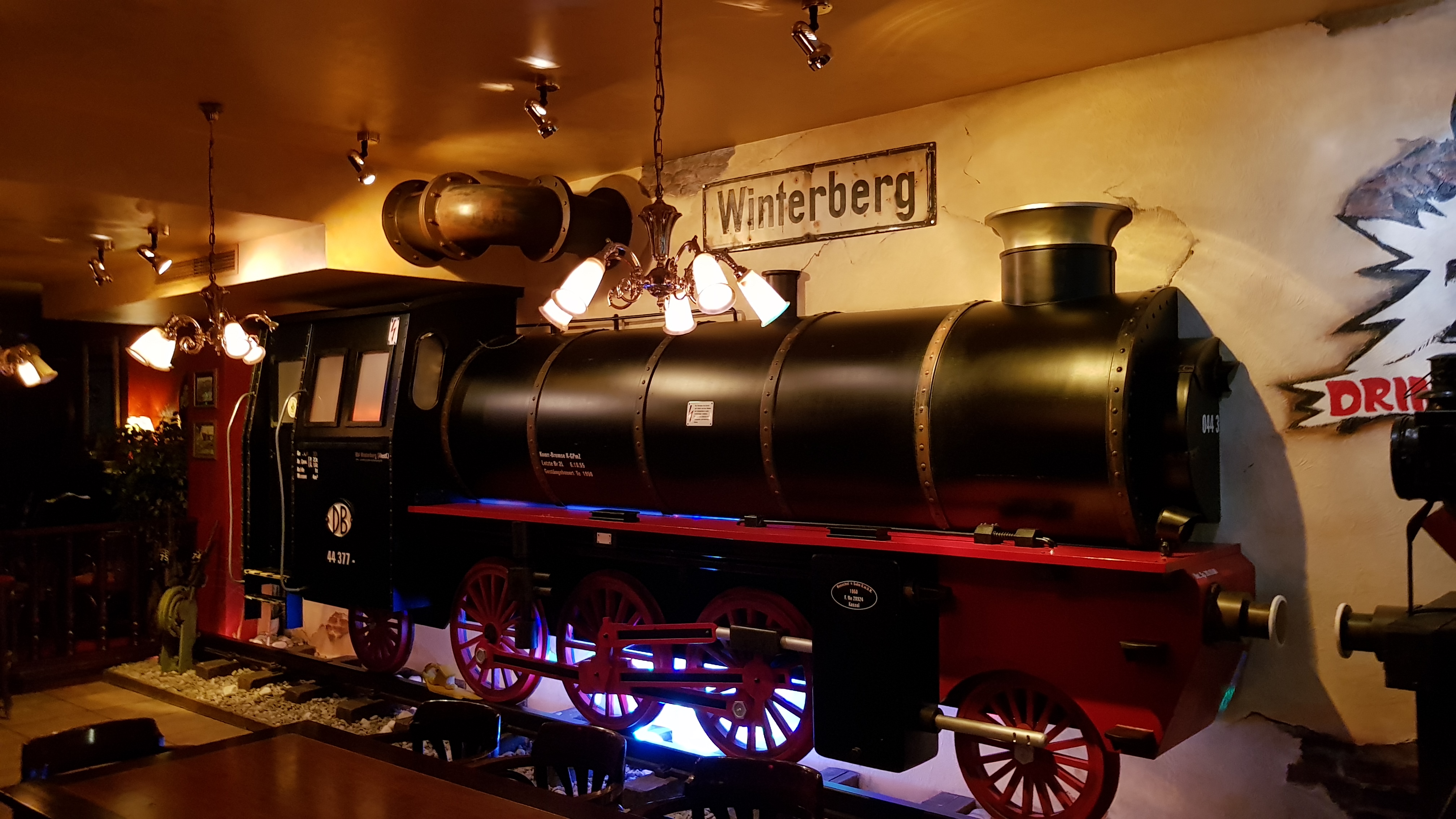 Eine Dampflok vor einer Wand, darüber ein Schild mit der Aufschrift Winterberg. Im Vordergrund hängen Lampen von der Decke.
