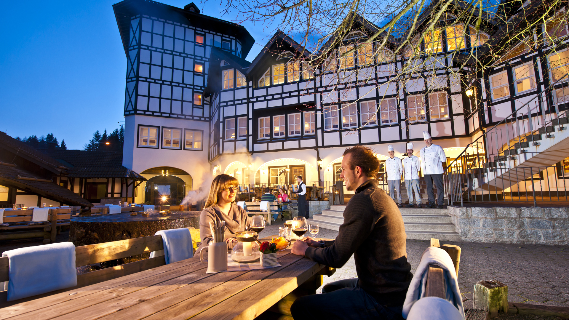 Zwei Personen beim Essen auf einer Außenterrasse mit einem Hotel im Fachwerkstil im Hintergrund.