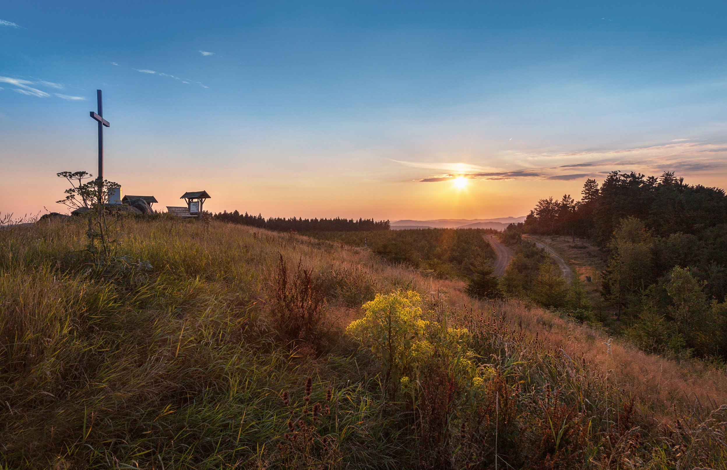 Ein Kreuz auf einem Hügel mit weitläufiger Aussicht und der aufgehenden Sonne im Hintergrund.