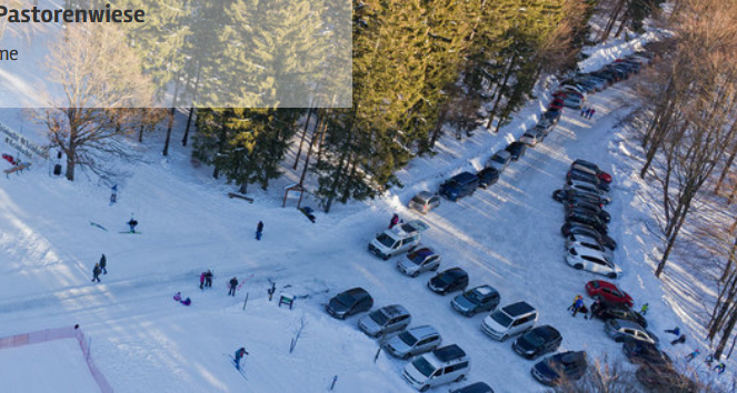 Drohnenaufnahme eines mit Autos beparkten, verschneiten Parkplatzes umgeben von Bäumen.