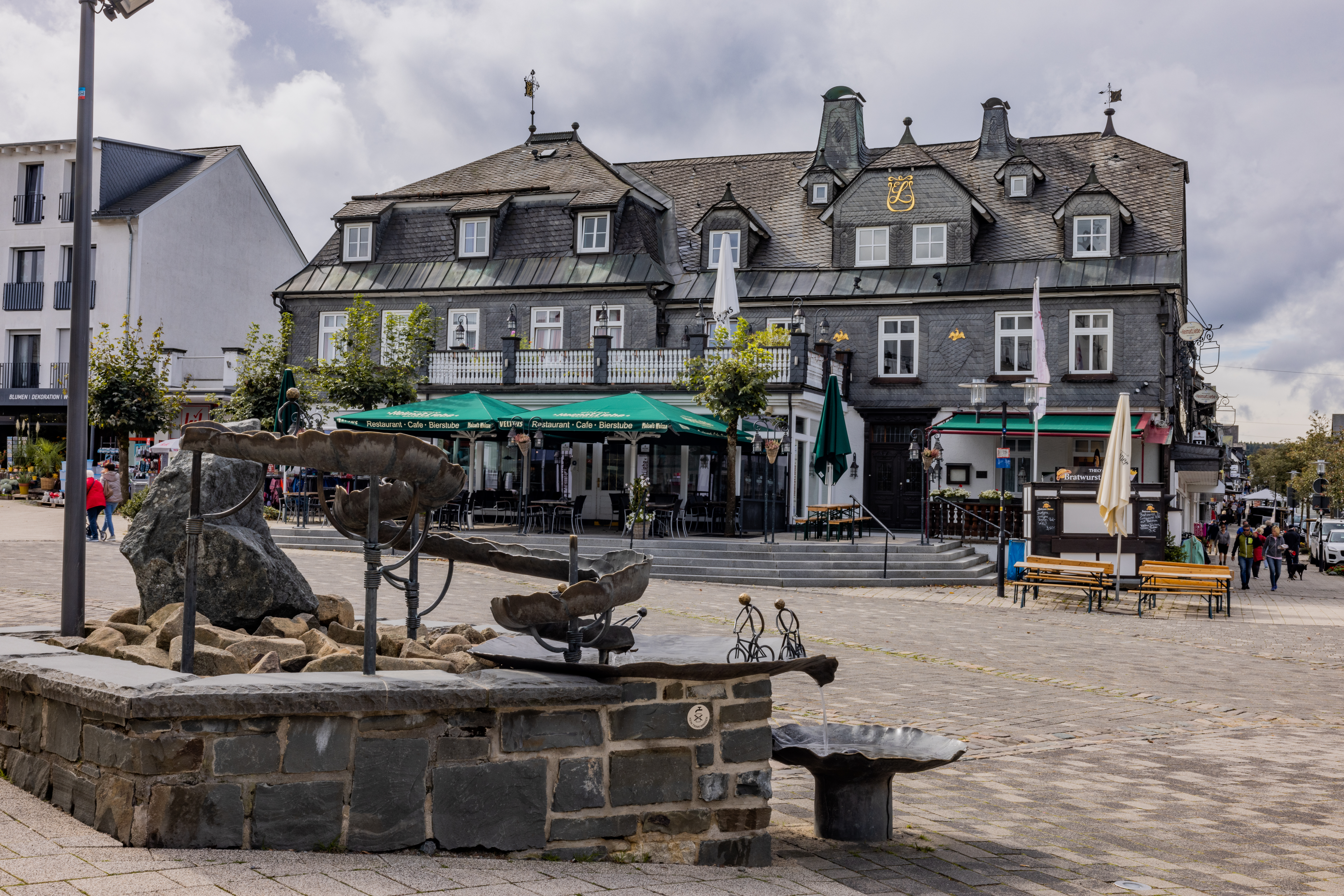 Ein Brunnen mit einem Wasserlauf mit einem Marktplatz und einem verschieferten Gebäude im Hintergrund.
