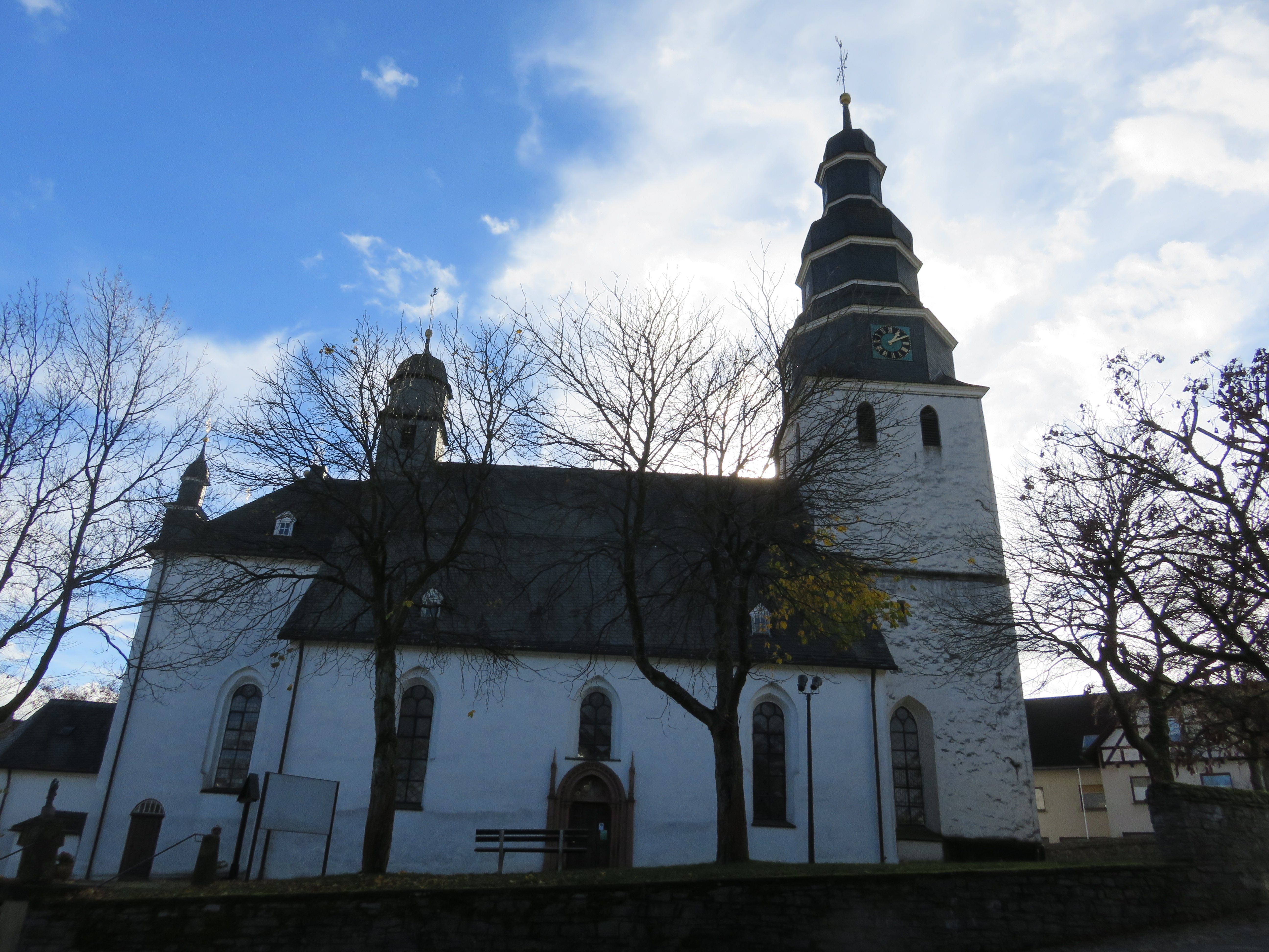 Eine Kirche umgeben von Bäumen vor blauem, bewölktem Himmel.