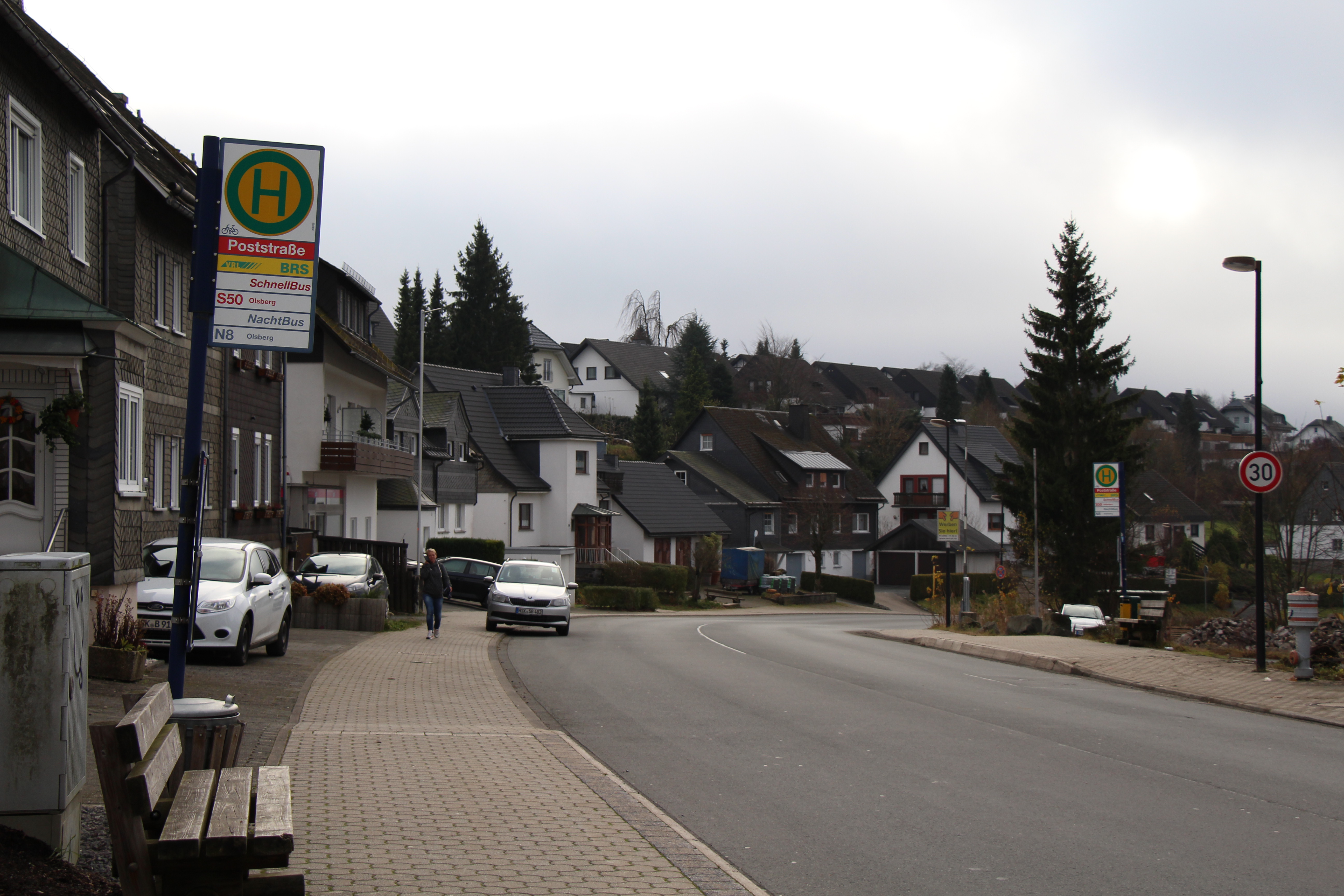 Ein Bushaltestellenschild neben einer Bank mit Häusern im Hintergrund.