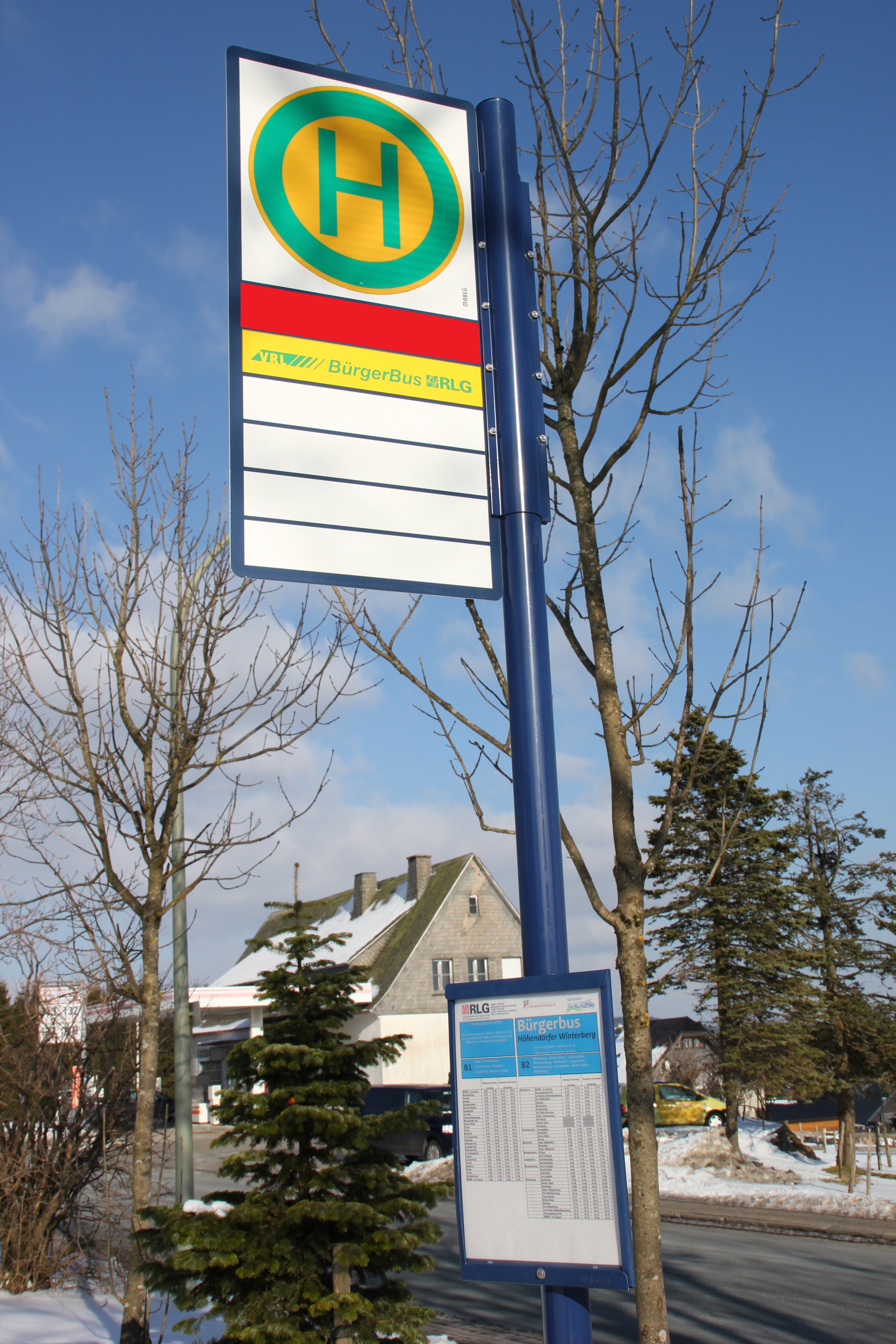 Ein Bushaltestellenschild mit Bäumen und einem Haus im Hintergrund.