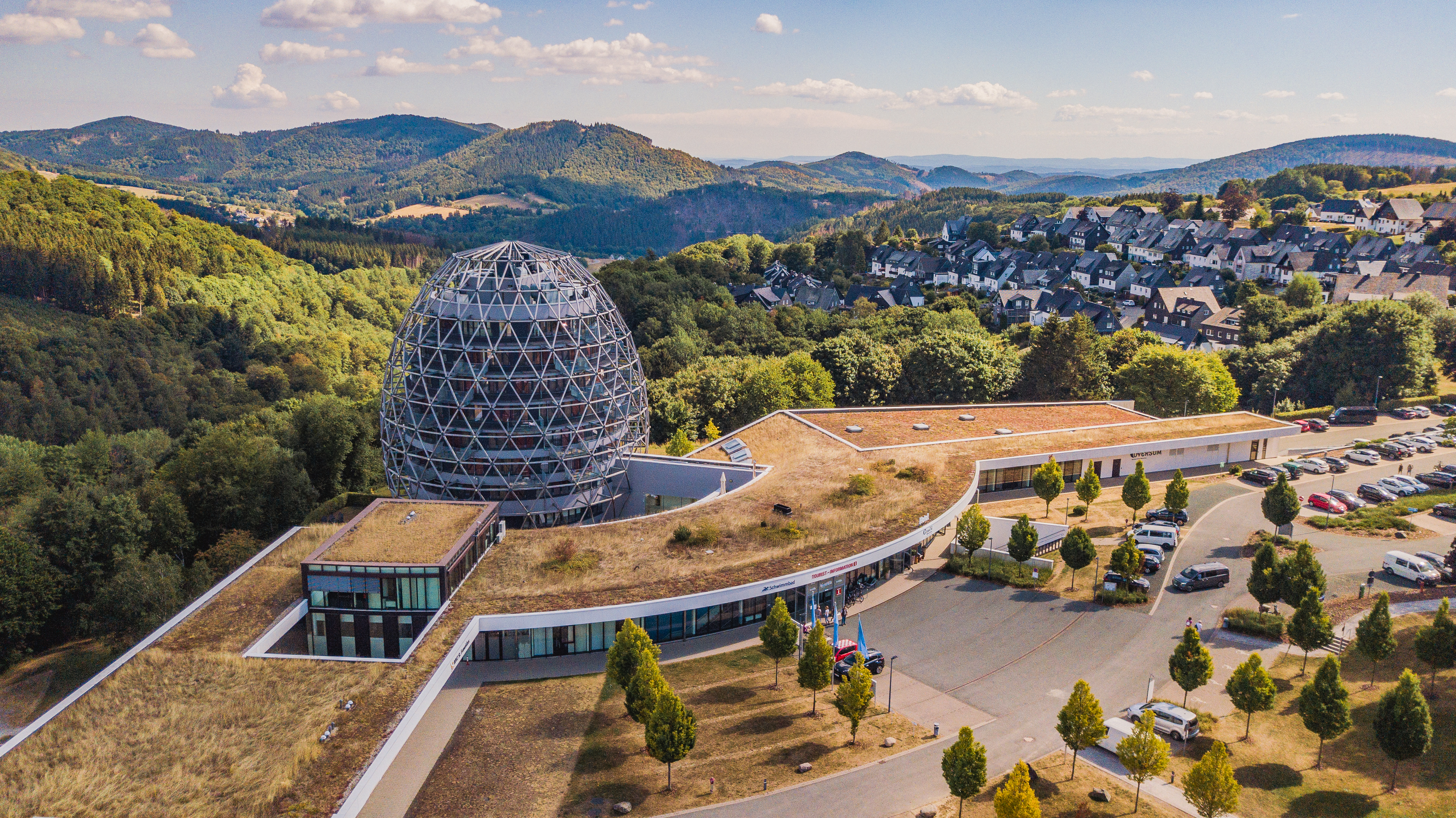 Drohnenaufnahme des Hotels Oversum mit der Tourist Information und dem Schwimmbad vor einer Berglandschaft.