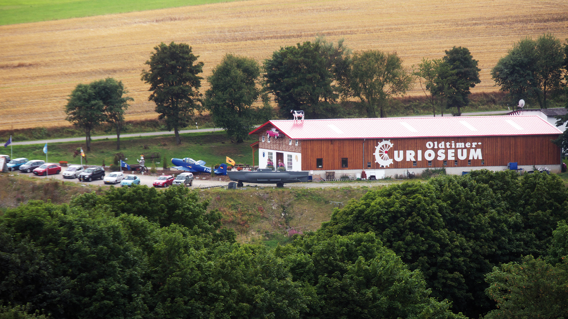 Ein Gebäude mit der Aufschrift Oldtimer Curioseum mit Parkplatz umgeben von Wiesen und Bäumen.