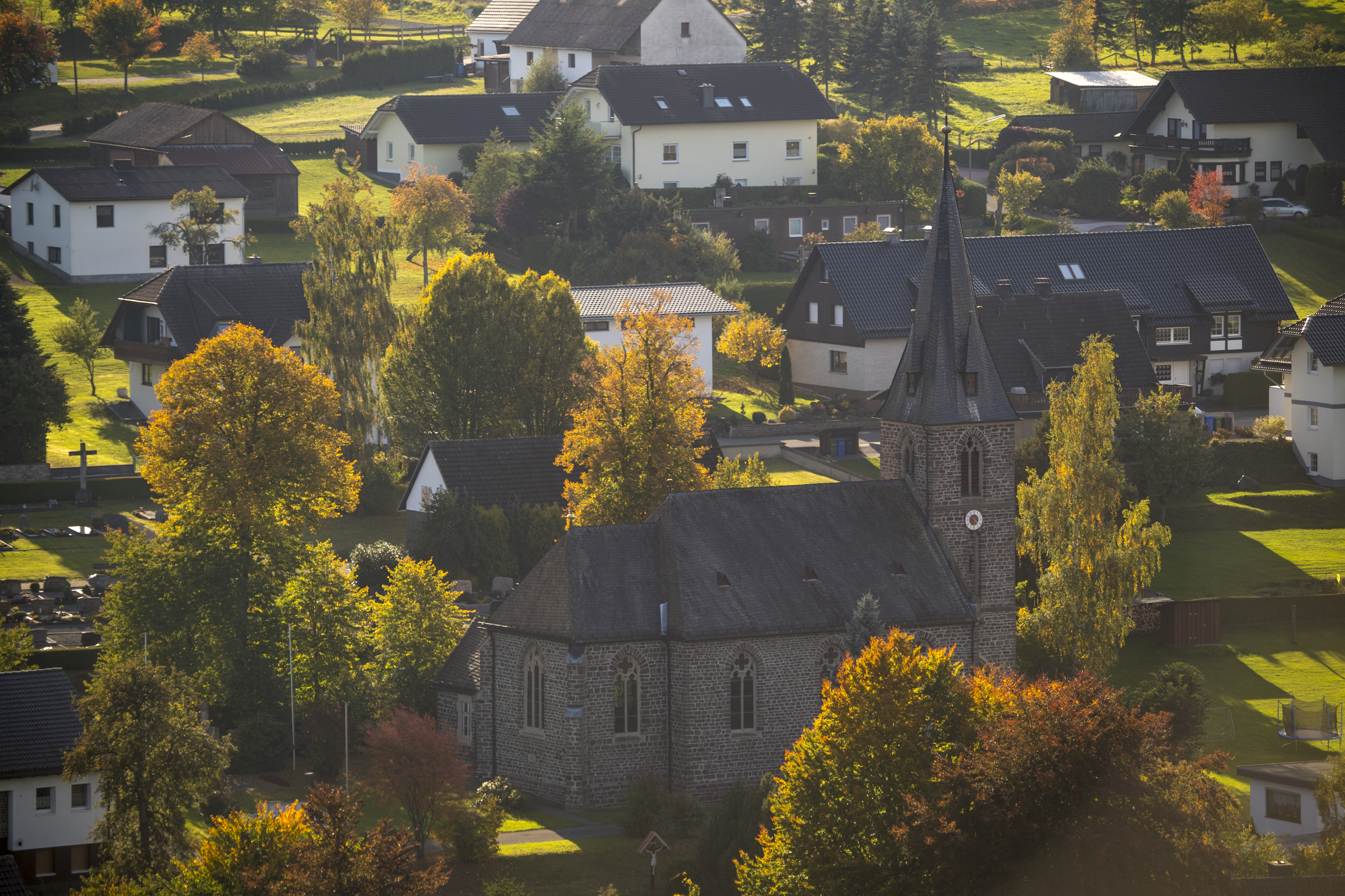 Drohnenaufnahme einer Kirche umgeben von einigen Wohnhäusern, Wiesen und Bäumen.